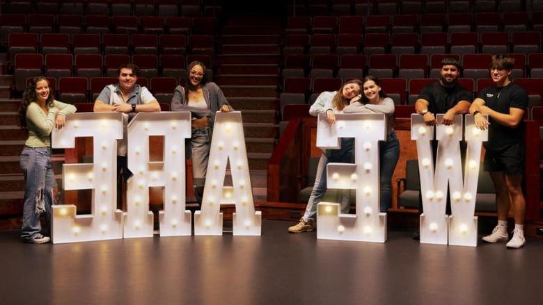 New Kensington students standing behind large letters that spell "WE ARE" inside the campus theatre.